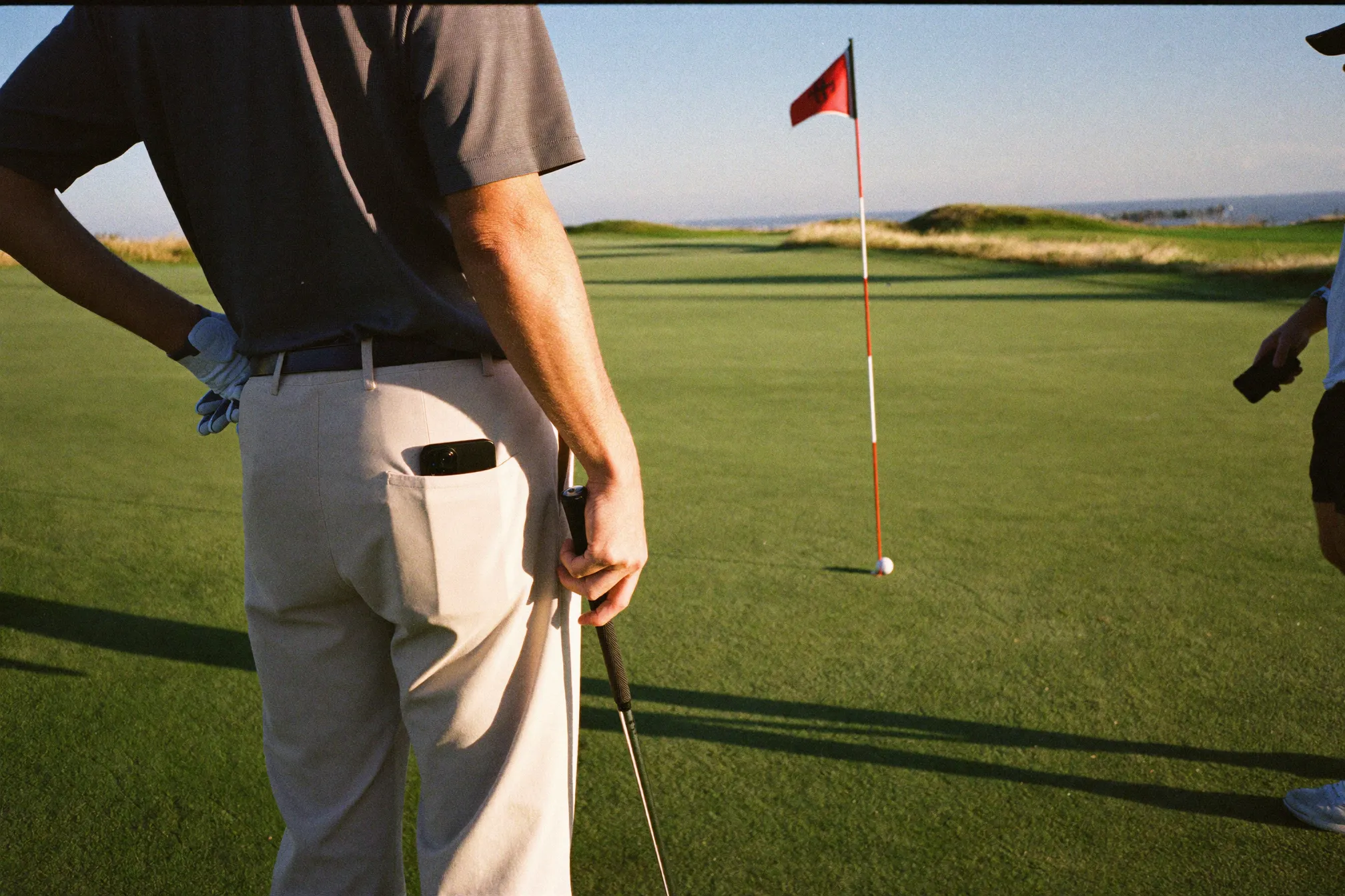 A golfer standing on a green at golden hour with the flag and the sea in the distance
